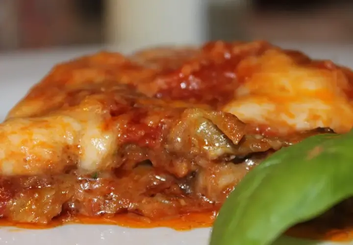 Rebanada de parmigiana di melanzane, con capas de berenjena, salsa de tomate y queso derretido, adornada con una hoja de albahaca fresca.
