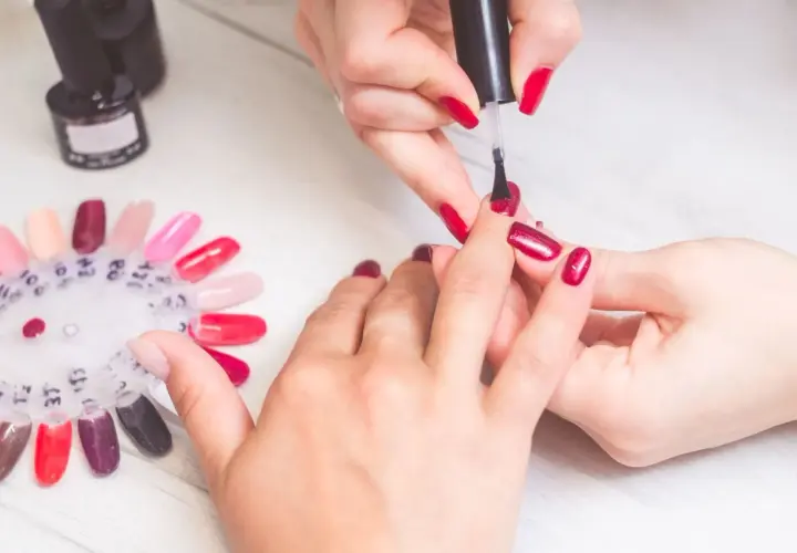 Krok po kroku: jak zrobić manicure. Dłonie w trakcie malowania paznokci na czerwono, obok paleta kolorów lakierów.