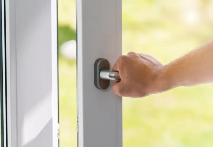 Dłoń chwyta metalową gałkę drzwi, otwierając je na zielony ogród.