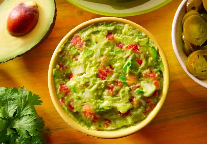 Miseczka guacamole z awokado, pomidorami i cebulą, obok połówka awokado i plasterki jalapeño.