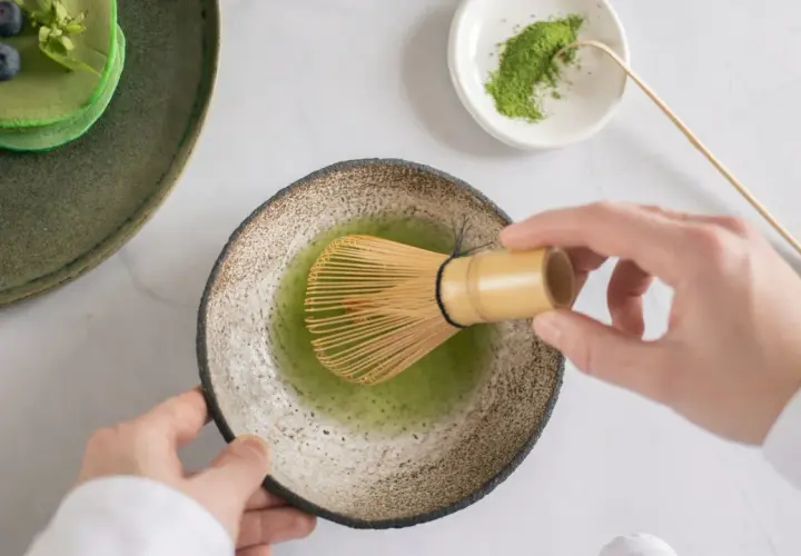 Matcha-Zeit: Hände bereiten grünen Tee mit einem Chasen zu. Daneben ein Stapel grüner Pfannkuchen mit Blaubeeren.