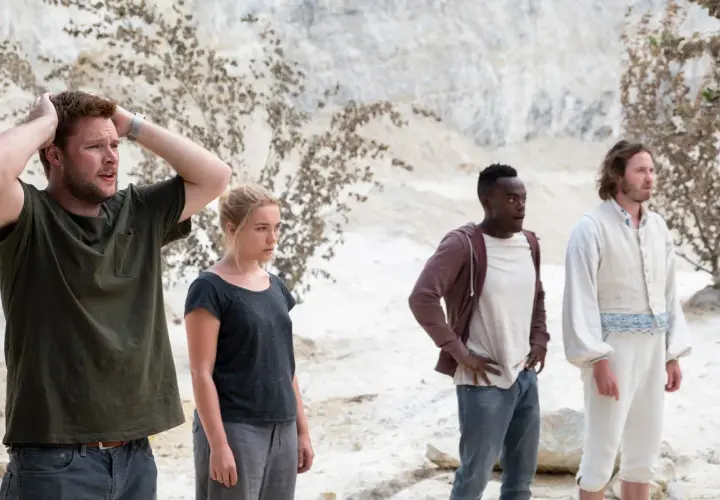 Fyra personer står i en öde, ljus miljö. En man med händerna i håret, en kvinna med blicken fäst framåt, en man med öppen mun och en man i vit, gammaldags klädsel. Scen från filmen Midsommar.