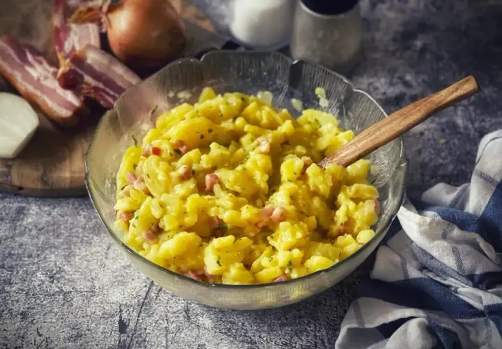 Schüssel mit warmem Kartoffelsalat, garniert mit Speck und Zwiebeln. Ein Holzlöffel liegt darin.