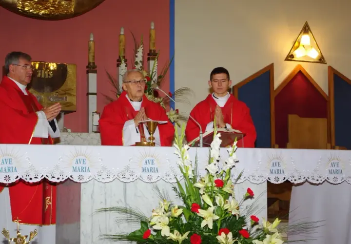 Trzech księży w czerwonych szatach podczas mszy, przed ołtarzem z kwiatami i napisem "Ave Maria".
