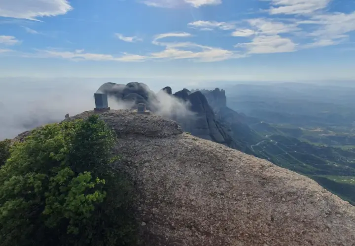 Cumbre rocosa con un monolito en Sant Jeroni, Montserrat. Nubes y montañas se extienden hasta el horizonte.