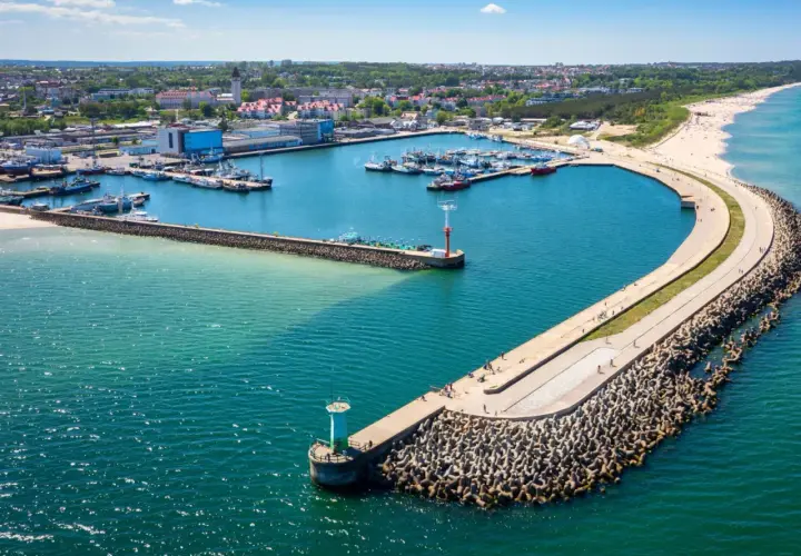 Port w Władysławowie z licznymi łodziami i falochronem, na którym widać spacerujących ludzi. W oddali plaża i zabudowania.