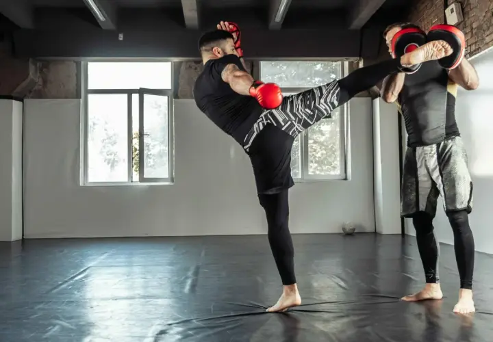 Trener łapie kopnięcie low kick. Dwóch mężczyzn w rękawicach treningowych ćwiczy sztuki walki na sali.