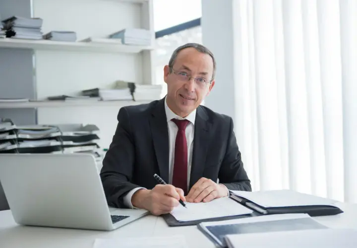Mężczyzna w garniturze podpisuje umowę najmu u notariusza. Na biurku laptop i dokumenty.