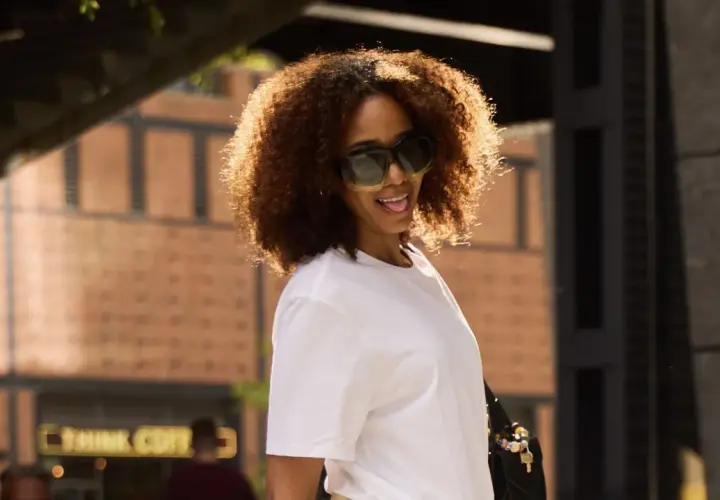 Frau mit lockigem Haar und Sonnenbrille trägt ein weißes T-Shirt.