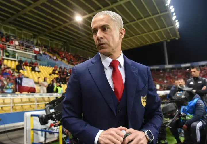 Sylvinho w granatowym garniturze i czerwonym krawacie, z emblematem na piersi, stoi na tle trybun stadionu.