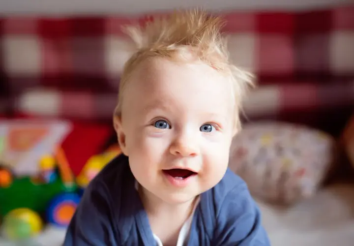 Uśmiechnięte niemowlę z blond czuprynką, któremu jakby nie rosną włosy. Ma niebieskie oczy i wesołe spojrzenie.