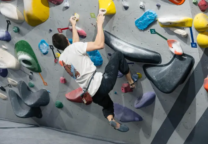 Mężczyzna w białej koszulce wykonuje trudny ruch podczas wspinaczki boulderingowej na kolorowej ściance.