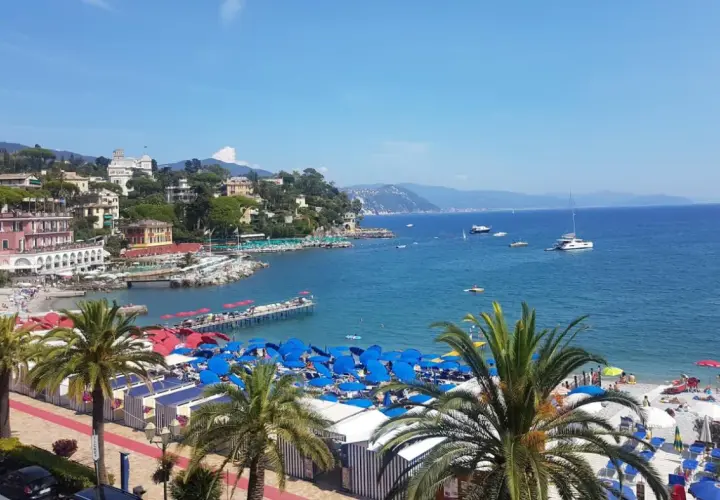 Plage animée à Santa Margherita Ligure, Italie, avec parasols bleus, cabines rayées, palmiers et bateaux sur la mer bleue.