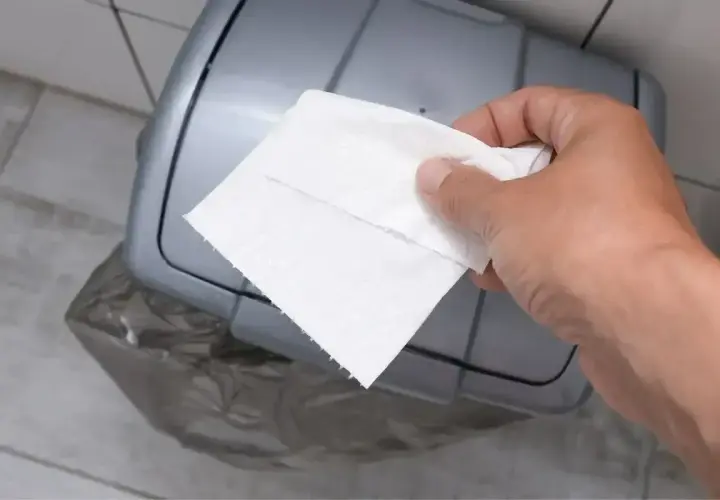Dłoń z ręcznikiem papierowym zbliża się do kosza. Właśnie tu, gdzie wyrzucać ręczniki papierowe, by zachować czystość.