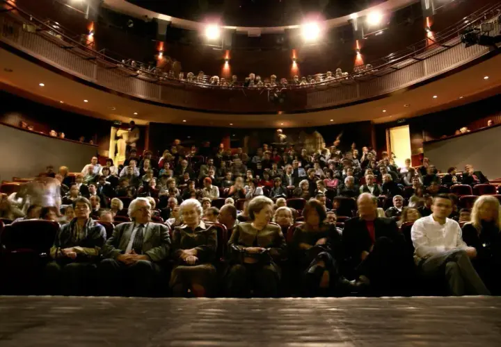 Widownia w teatrze, ludzie siedzą na miejscach, widać balkon z widzami.