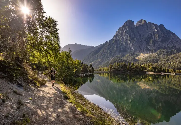 Senderista camina por el sendero junto al estany de Basturs, reflejando la majestuosa montaña y el bosque en sus aguas tranquilas.