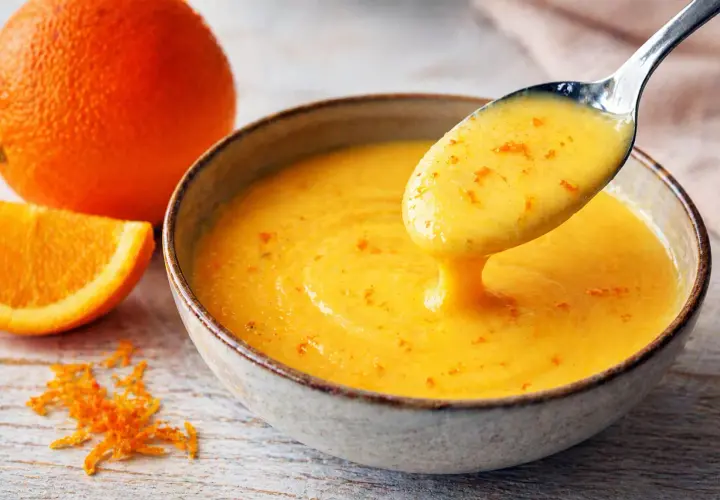Une cuillère dégouline d'une sauce maltaise onctueuse, parsemée de zestes d'orange. Une orange entière et une tranche sont à proximité.