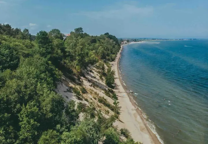 Mechelinki klif porośnięty drzewami, z piaszczystą plażą i turkusowym morzem. W oddali widać zabudowania i falochron.