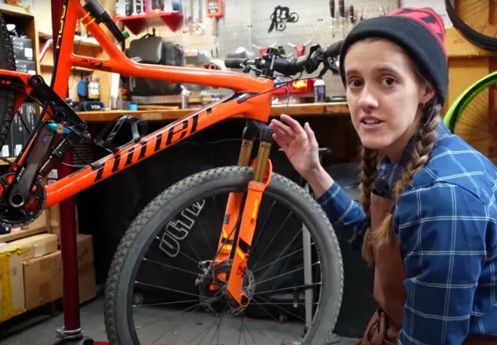 A woman in a blue plaid shirt and beanie explains bike maintenance, pointing to the orange frame and front suspension of a mountain bike.