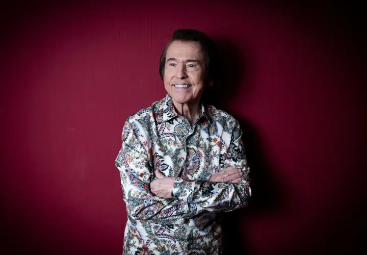 Raphael, sonriente y con los brazos cruzados, posa ante un fondo rojo. Su camisa estampada evoca la energía de sus conciertos.