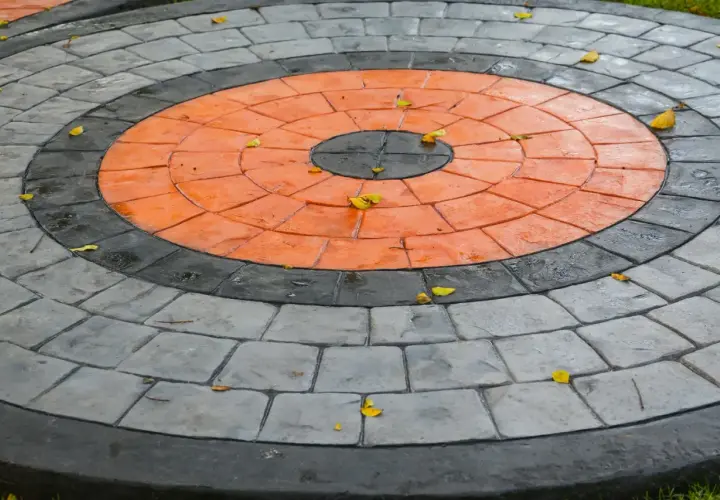 Kreisrunder Weg aus grau und orangefarbenen Pflastersteinen, die wie ein Ziel aussehen. Einige gelbe Blätter liegen darauf.
