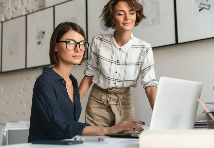 Dwie kobiety pracują przy laptopie. Jedna w okularach, druga stoi za nią. Ich współpraca to przykład numerologiczna 4 – stabilność i solidność.