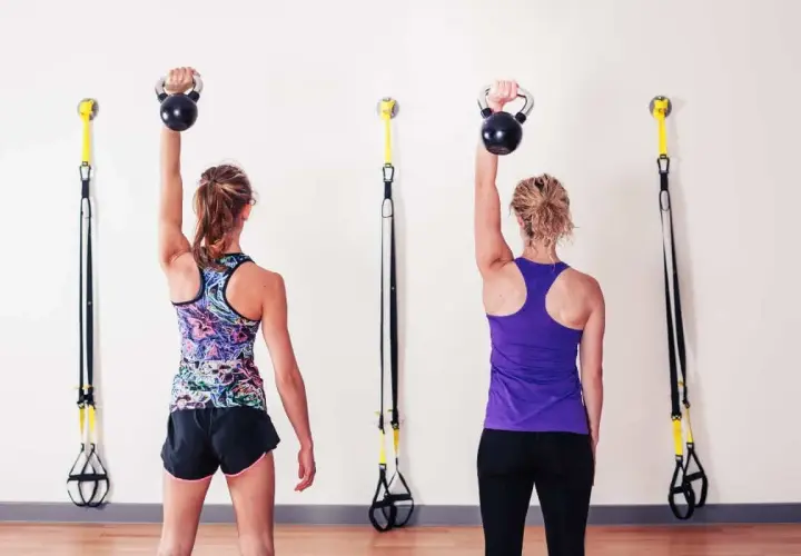 Dwie kobiety wykonują rzs ćwiczenia z kettlebellami. W tle wiszą taśmy TRX.