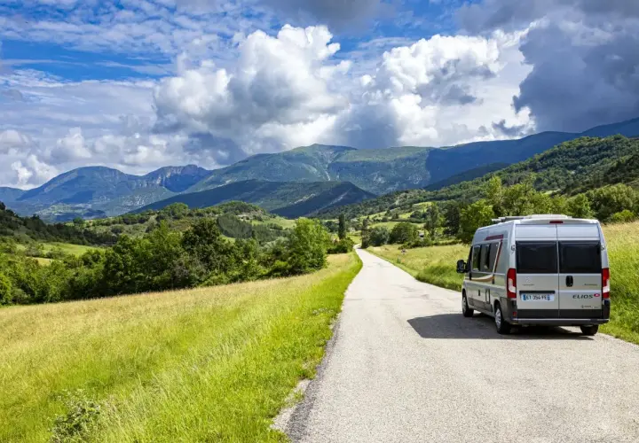 Un camping-car Elios sillonne une route sinueuse à travers les paysages verdoyants du Vercors, sous un ciel bleu parsemé de nuages.
