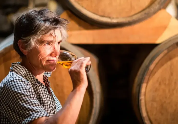 Une femme déguste un verre de vin de Pau, entourée de fûts de chêne dans une cave.