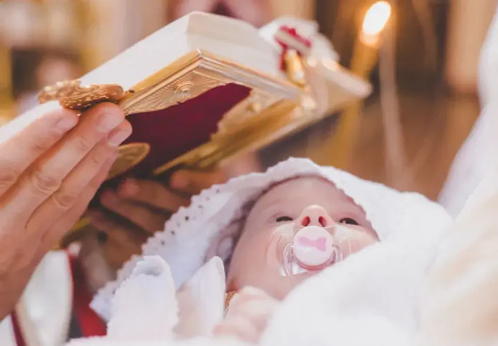 Dziecko z smoczkiem podczas ceremonii. Ksiądz trzyma księgę, a obok pali się świeca. Ile trwa chrzest?