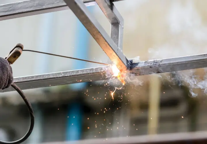 Ręka w rękawicy trzyma elektrodę, wykonując spawanie łukowe metalowych profili. Iskry i dym unoszą się w powietrzu.