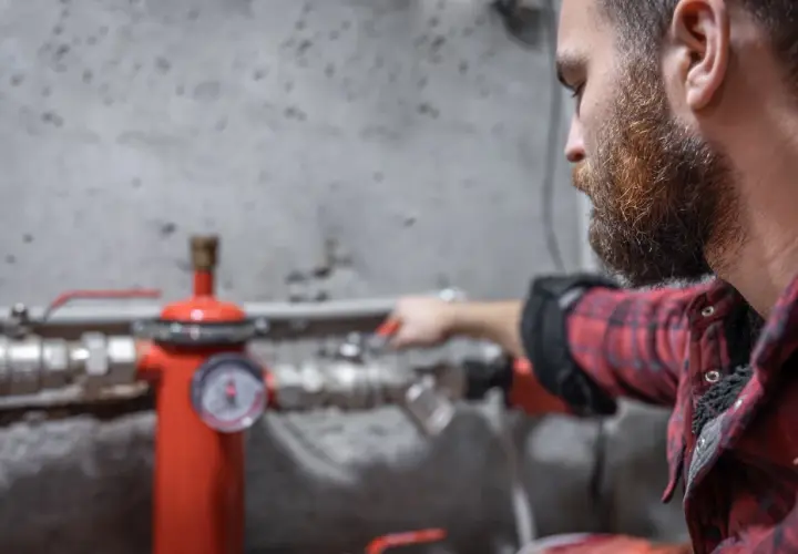Uomo con barba controlla un manometro su un tubo rosso. Sento rumore nei tubi dell'acqua, forse un problema da risolvere.