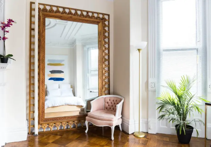 A grand pier mirror with an ornate gold frame reflects a bed and abstract art. A pink velvet chair sits beside it.