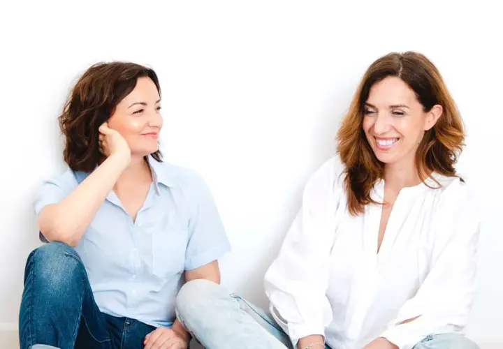 Dos mujeres sentadas, una con camisa azul y jeans, la otra con blusa blanca. Parecen compartir un momento laconicum, de pocas palabras pero profunda conexión.
