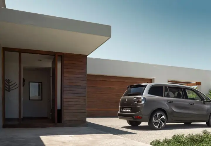 Szary minivan Citroën C4 Picasso zaparkowany przed nowoczesnym domem z drewnianą elewacją.