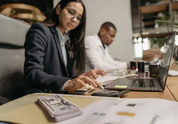 Kobieta w okularach liczy na kalkulatorze, obok stos banknotów i laptop.