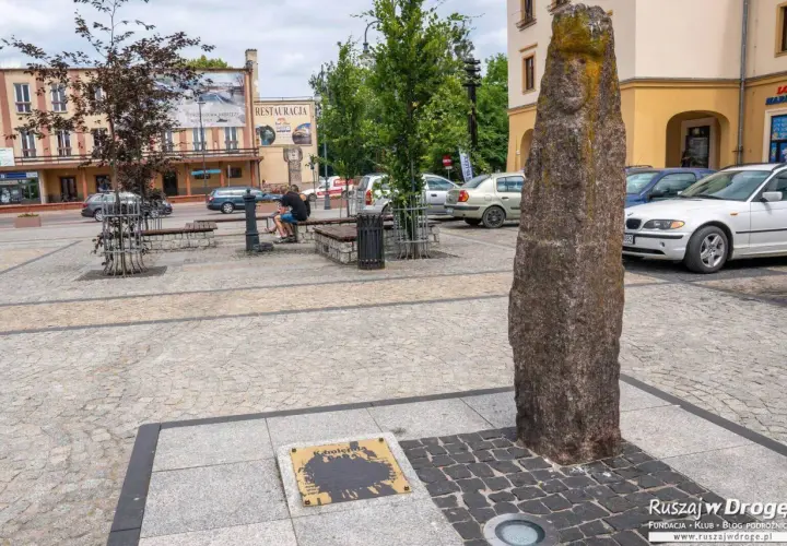 Kamienny pomnik i tablica z napisem "Kamienna" to ciekawe atrakcje na rynku. Obok ławki i drzewa, w tle budynki i samochody.