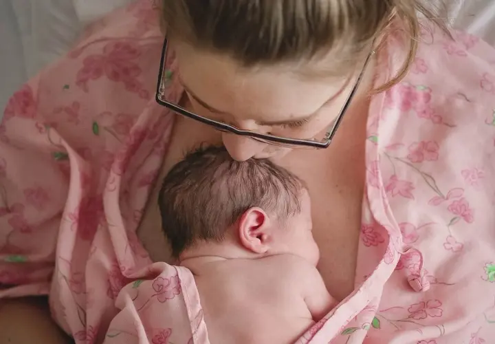 Mama całuje główkę noworodka, otulając go w różowy materiał. To piękny moment kangurowania, budowania więzi.