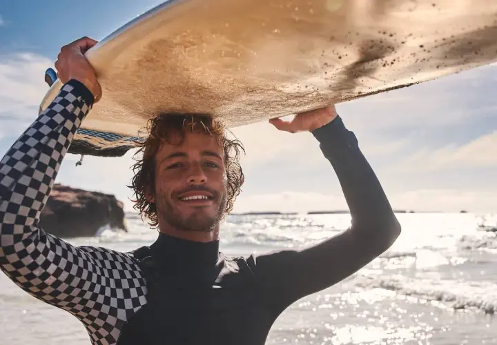 Lächelnder Surfer mit lockiger surfer frisur trägt Neoprenanzug und hält Surfbrett über dem Kopf.