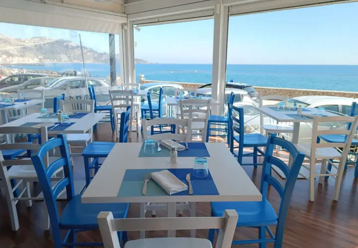 Tavoli apparecchiati con vista mare, perfetti per un apericena a Giardini Naxos. Sedie bianche e blu, un'atmosfera rilassante.
