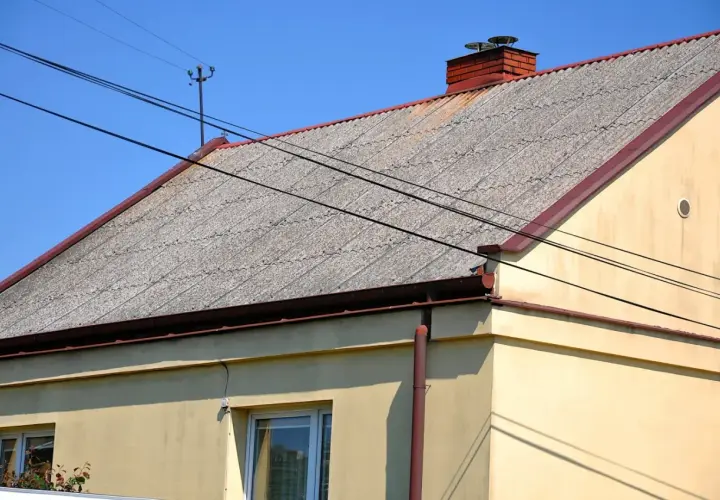 Dach pokryty falistym eternitem, z ceglanym kominem i liniami energetycznymi przecinającymi błękitne niebo.