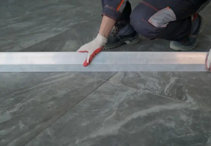 Mano con guanto bianco e rosso posiziona un profilo metallico su piastrelle grigie 60x120. Preventivo prezzo posa piastrelle 60x120.