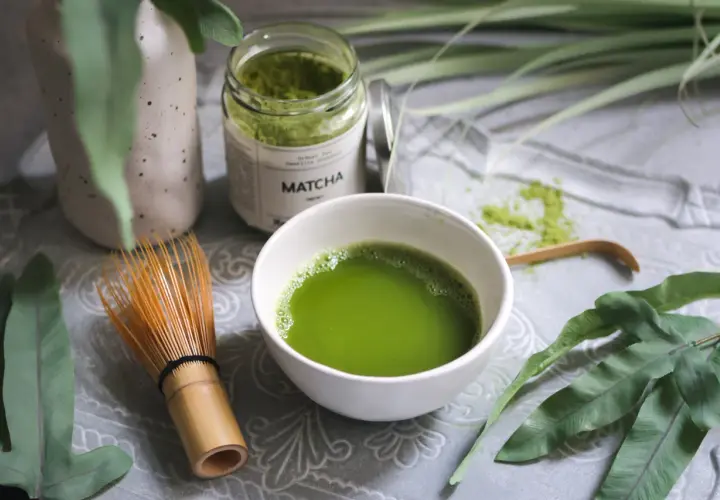 Matcha-Pulver in einem Glas, eine Schale mit zubereitetem Matcha und ein Bambusbesen. Die Matcha-Wirkung auf den Darm wird oft positiv beschrieben.