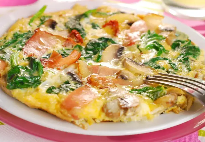 Omlet z szpinakiem, pieczarkami i bekonem na białym talerzu, widelcem.