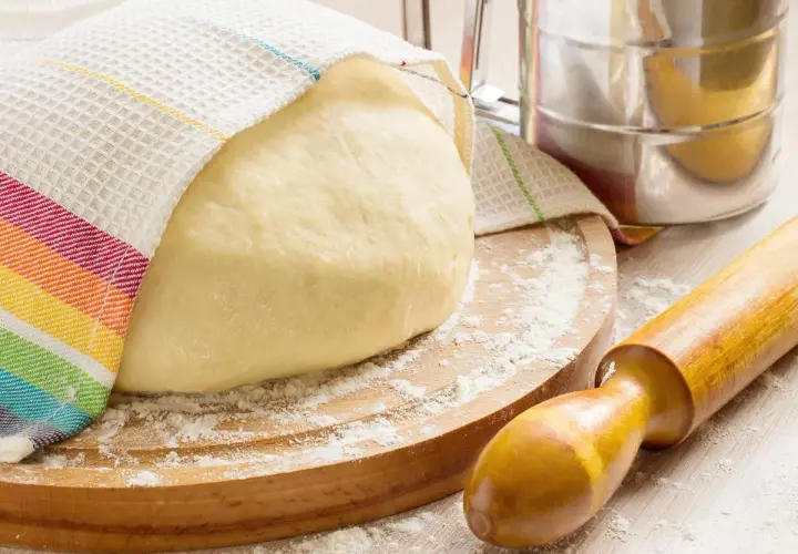 Masa de pan cubierta con un paño a rayas, sobre una tabla de madera con harina esparcida. Rodillo y tamizador de harina, ingredientes para hacer pan.