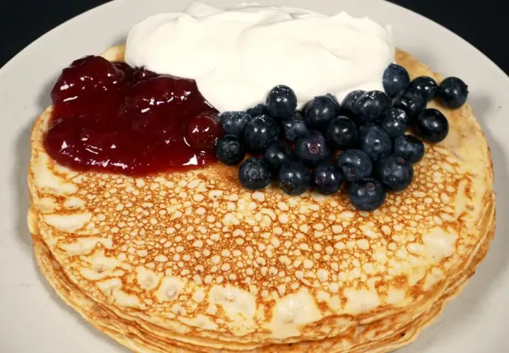 En hög med glutenfria pannkakor toppas med sylt, blåbär och grädde.