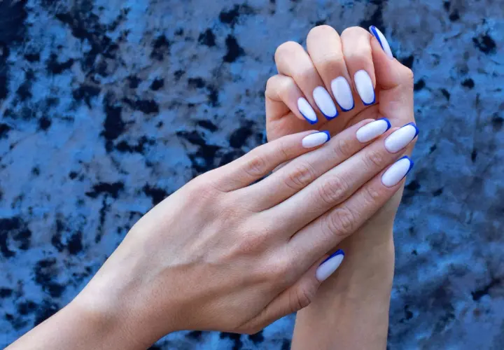 Eleganckie, białe paznokcie z granatowym french manicure, prezentowane na tle niebieskiego, aksamitnego materiału.