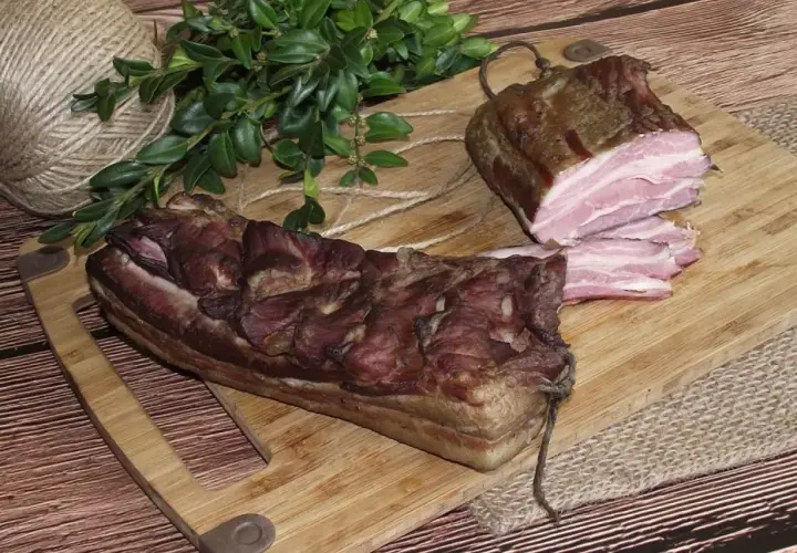 Dwa kawałki wędzonego boczku, jeden pokrojony w plastry, leżą na desce. Obok gałązka zieleni i sznurek.