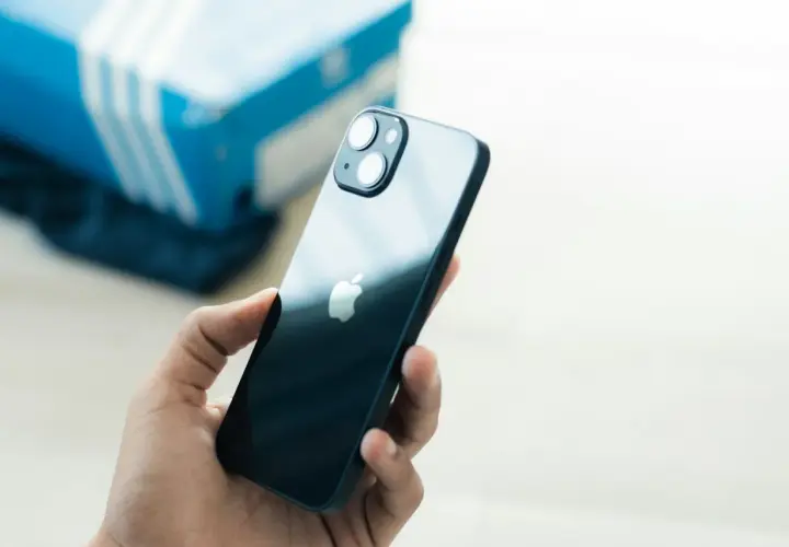 Dłoń trzyma ciemny iPhone 13, zastanawiając się, czy iPhone 13 jest wodoszczelny. W tle niebieskie pudełko.