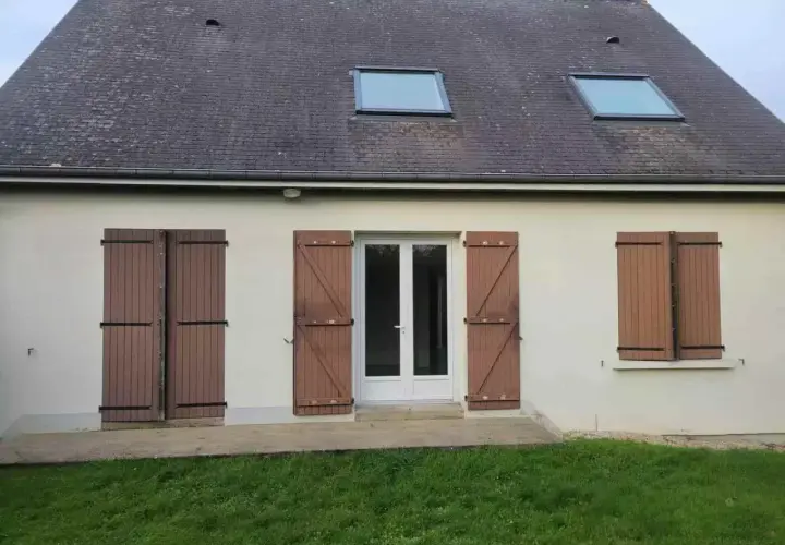 Façade d'une maison année 80, caractéristiques : toit en ardoise, deux fenêtres de toit, porte-fenêtre blanche et volets en bois marron.